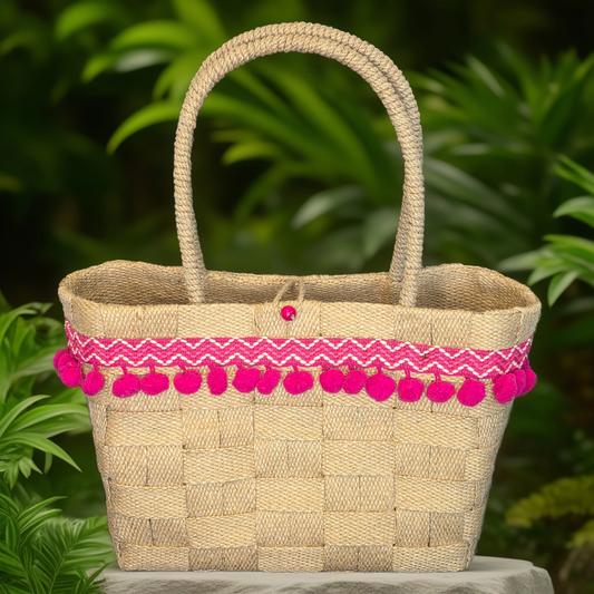 A beige jute basket handbag with pink pom pom trim, displayed against a tropical background with greenery.